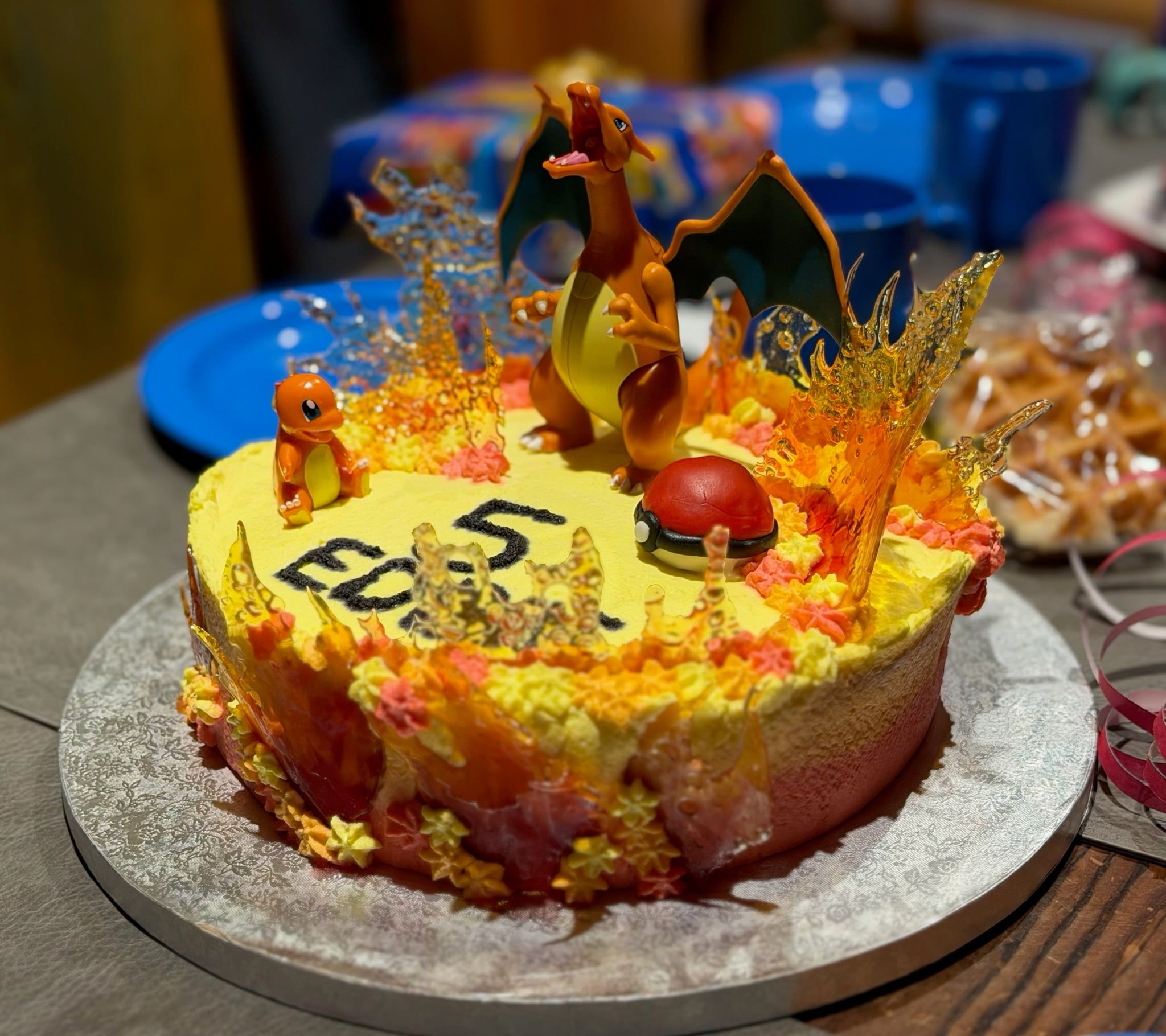 Gelb eingestrichene Torte mit einer Glurak-Figur, Flammen aus Zuckerglas, einem Pokéball und einer kleinen Glumanda-Figur; vorn stehen ein Name und die Zahl Fünf in dunkler Schrift.