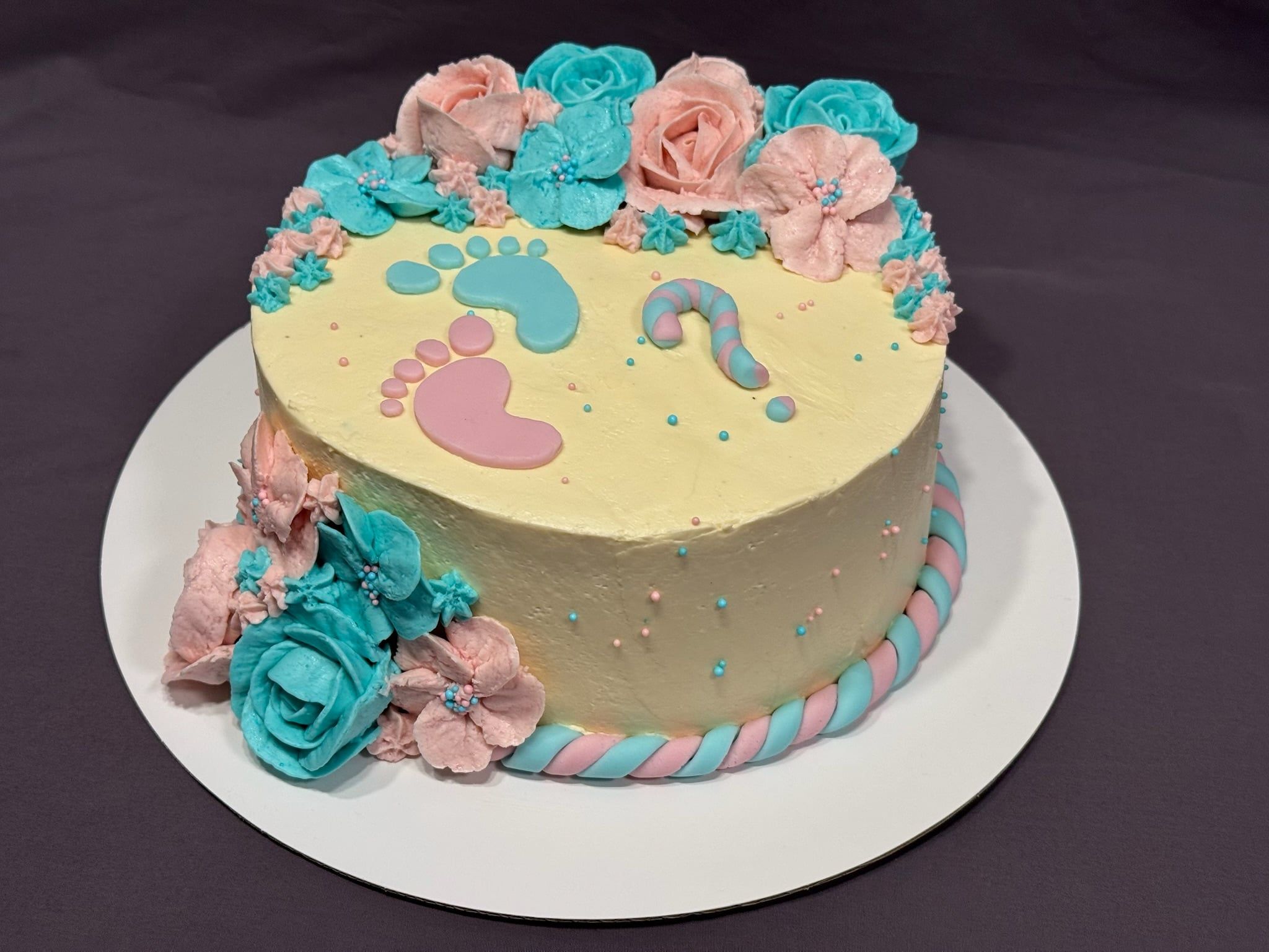 Runde Torte in Vanillegelb mit zwei Fußabdrücken in Rosa und Blau, einem gestreiften Fragezeichen und einem Kranz aus Buttercremeblüten.