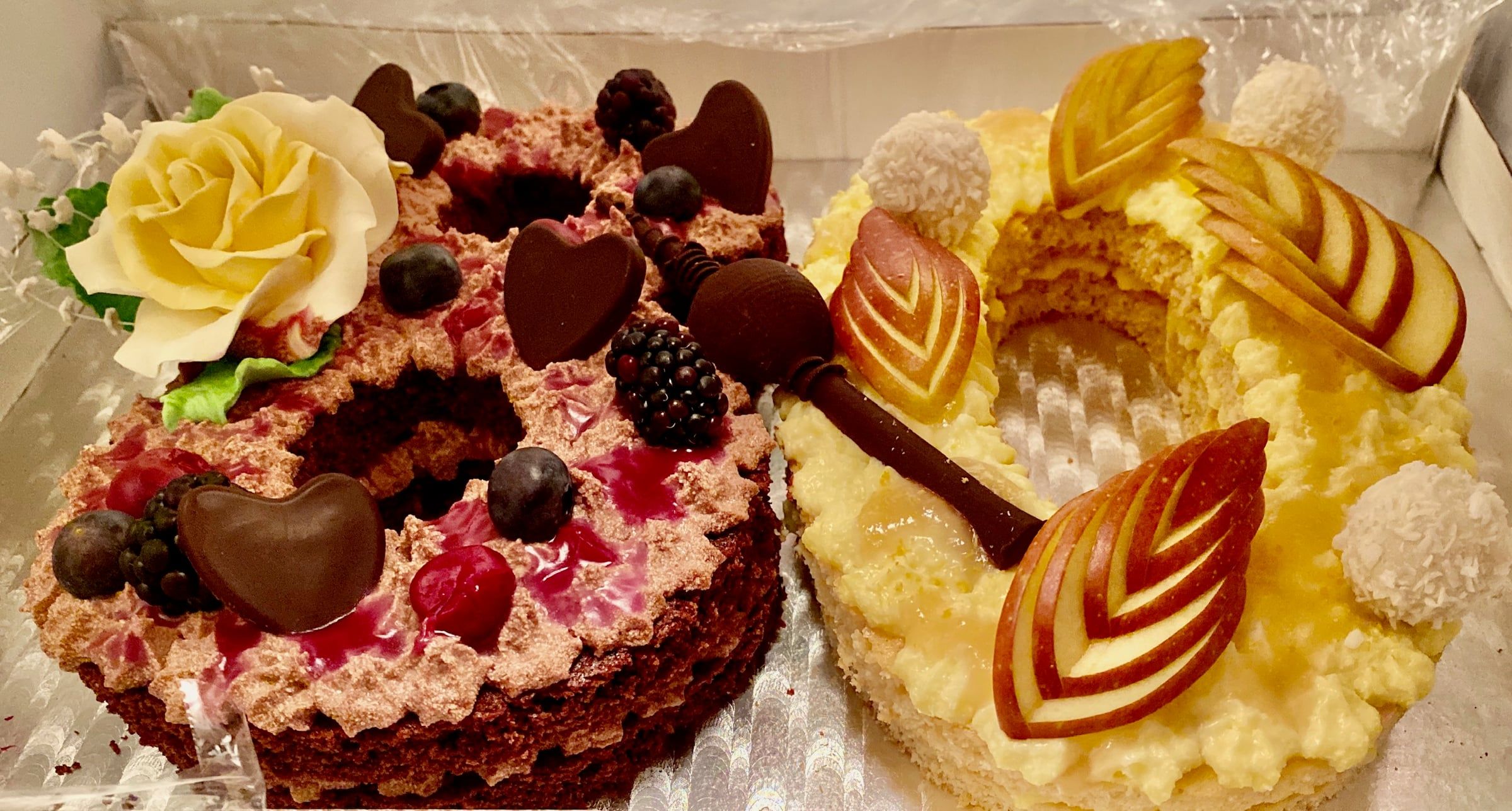Zwei Zahlentorten nebeneinander in der Transportbox, links eine dunkle mit Beeren, Schokoherzen und einer weißen Zuckerrose, rechts eine helle mit Apfelscheiben und Kokoskugeln.