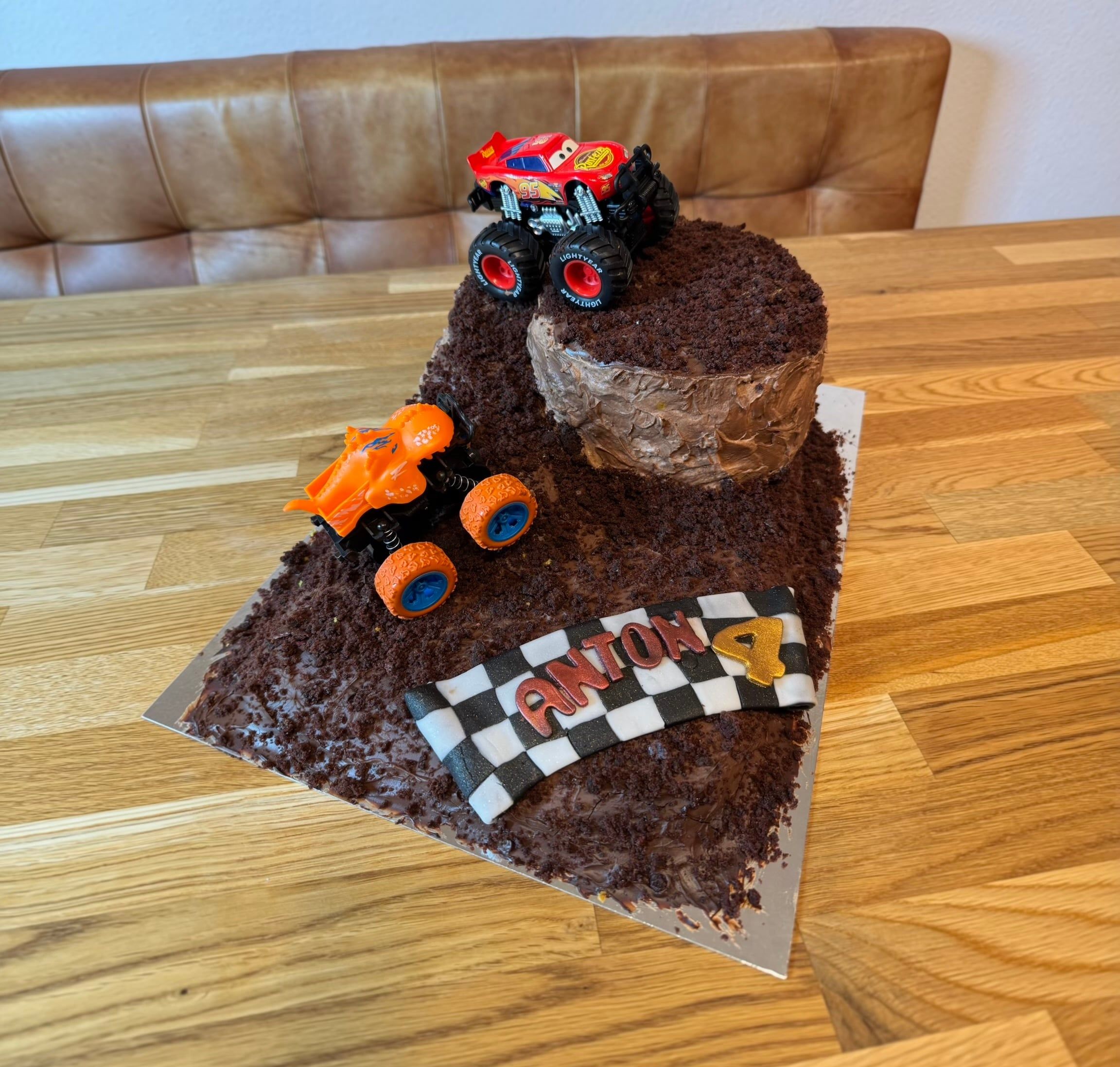 Zweistöckiger Schokoladenkuchen, überzogen mit Schokostreuseln, darauf zwei Monstertruck-Figuren und eine Zielflagge mit einem Namen und der Zahl Vier.