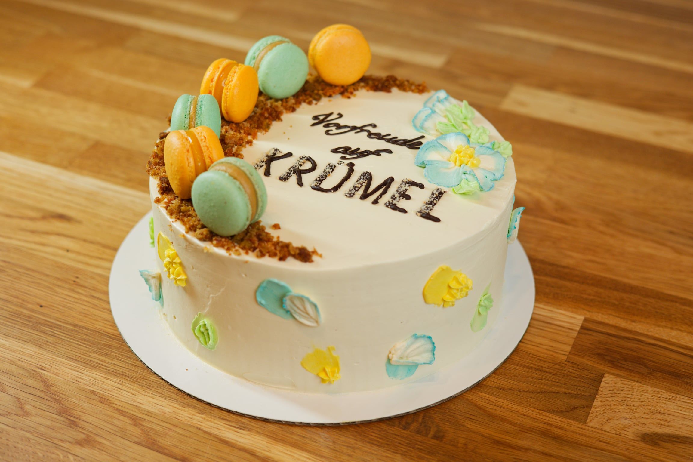 Runde Torte mit weißer Buttercreme, oben der Schriftzug „Vorfreude auf Krümel“ in dunkler Schokolade, dazu Macarons in Mint und Orange, Streusel und aufgemalte Blüten.
