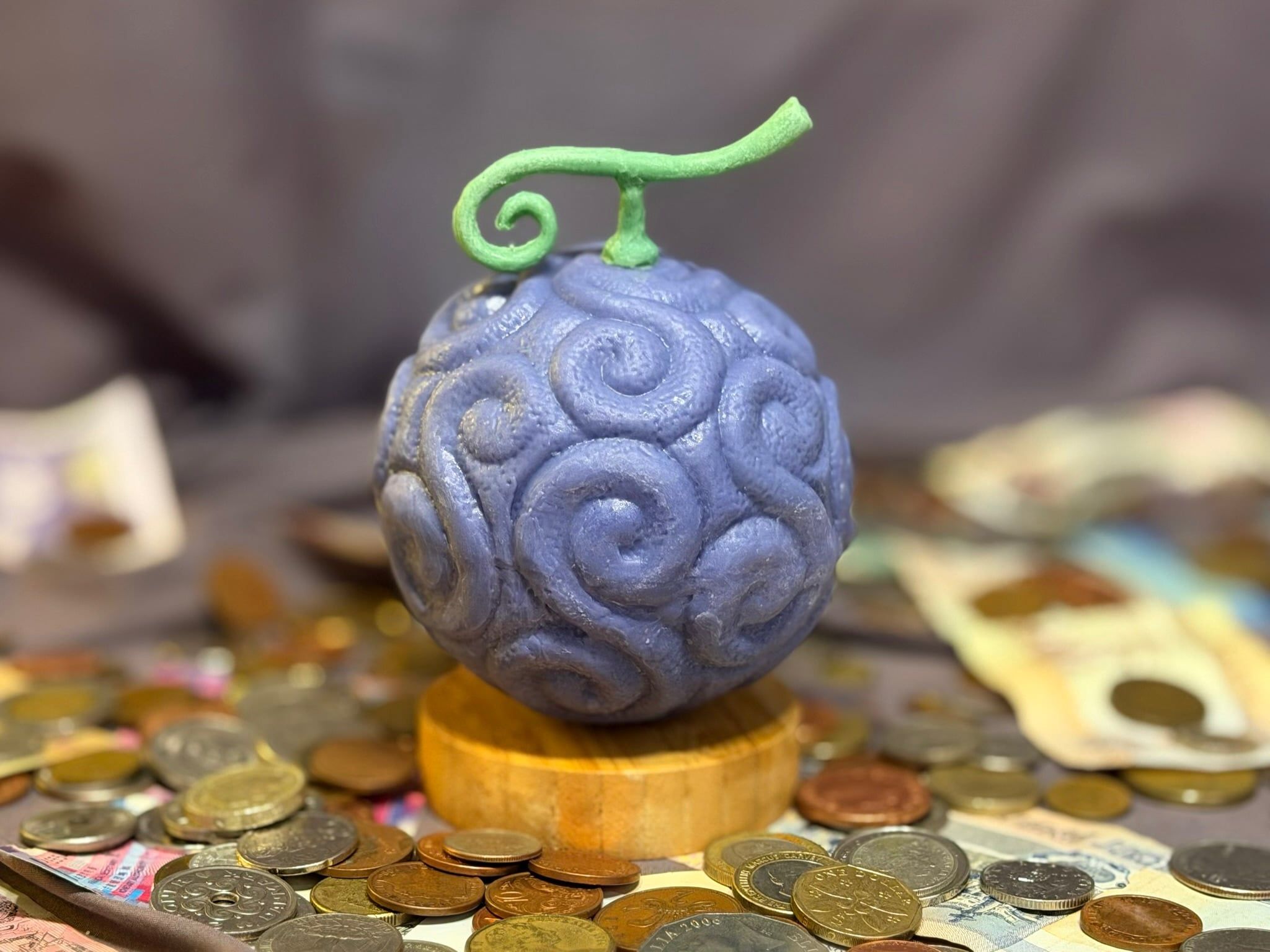 Eine violette Frucht aus Schokolade mit Spiralmuster und grünem Stiel, auf einem Holzsockel zwischen Münzen und Geldscheinen.