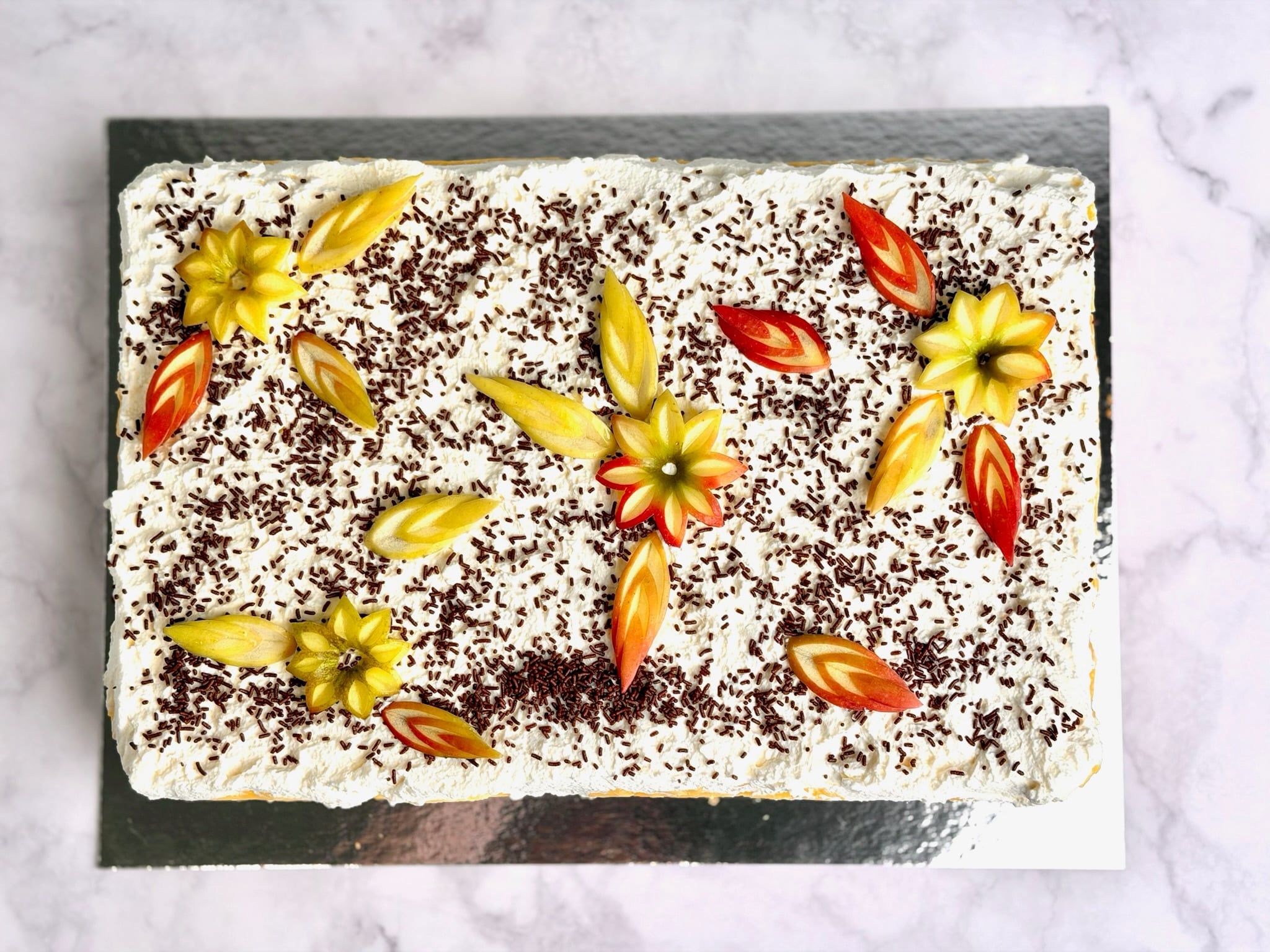 Dieselbe Torte von oben: Schokoraspeln über der Sahne, dazwischen Blüten aus Apfelscheiben in Gelb und Rot.