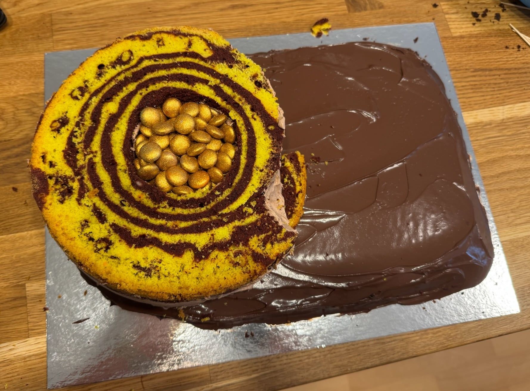 Derselbe Kuchen im Bau: ein angeschnittener runder Boden mit geringeltem Muster und goldenen Schokolinsen in der Mitte, daneben die mit Schokolade überzogene Platte.