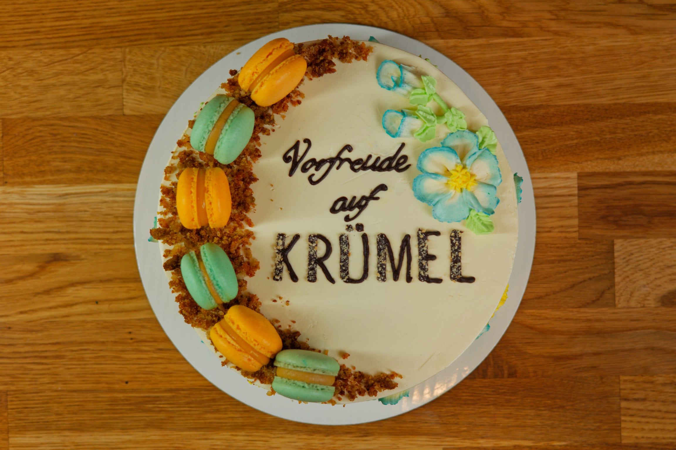 Dieselbe Torte von oben: ein Halbkranz aus Macarons und Streuseln, daneben der Schriftzug und eine blau-gelbe Blüte.
